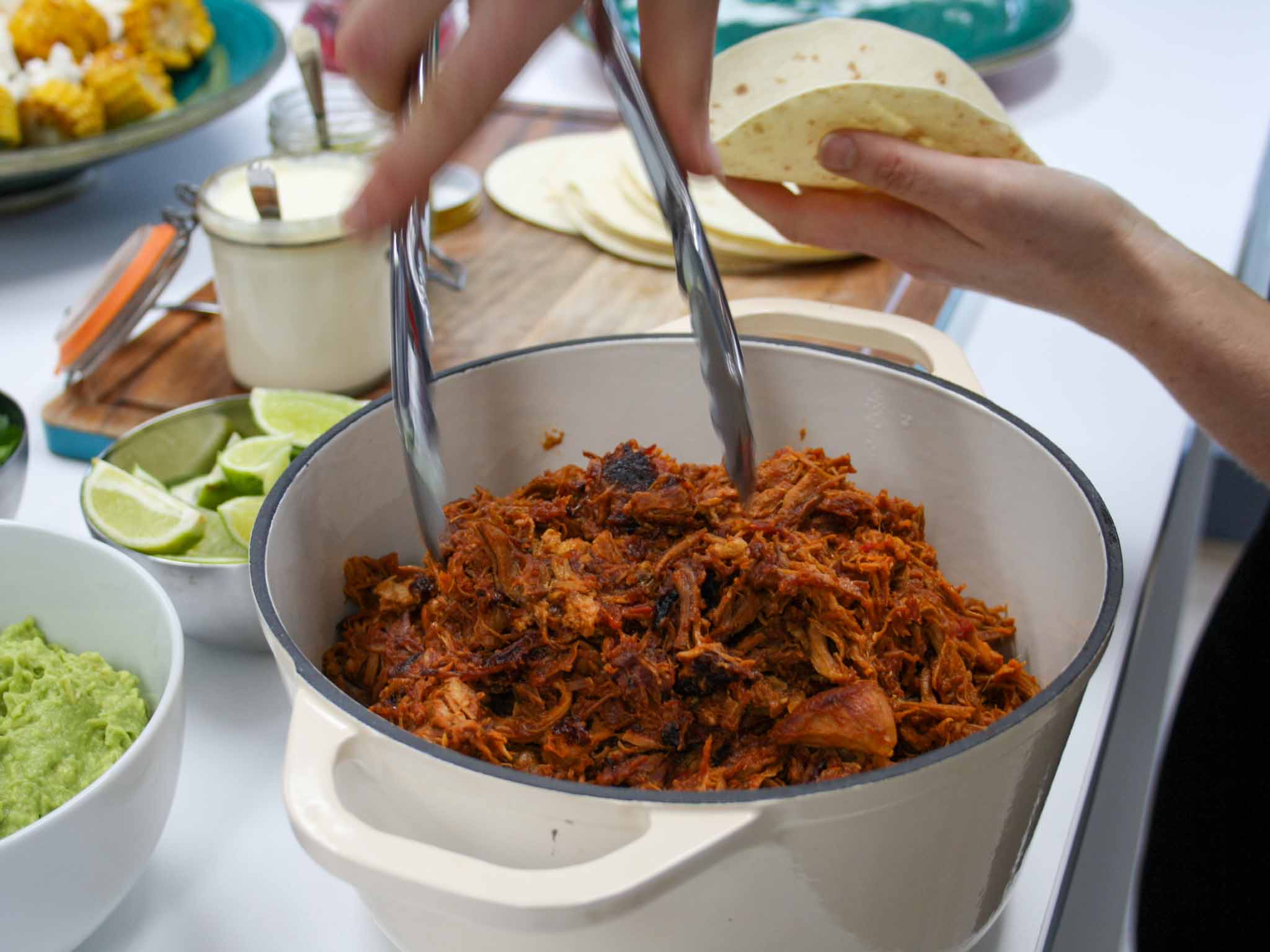 Close up of pot of pulled pork with tongs reaching in. Limes, tortillas, guacamole and sour cream in background.